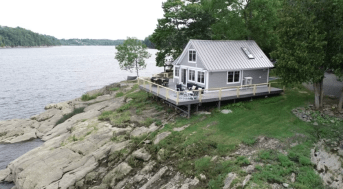 A cozy gray house with a deck overlooking a calm lake, surrounded by trees and rocky shoreline.