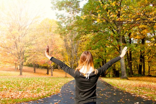A person with long hair stands with arms outstretched on a path surrounded by colorful autumn trees.