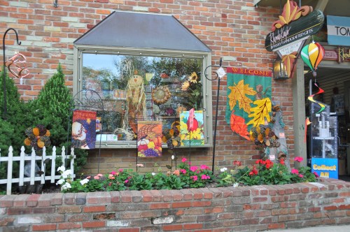 A charming storefront decorated with autumn leaves, flowers, and a "Welcome" sign, set against a brick wall.