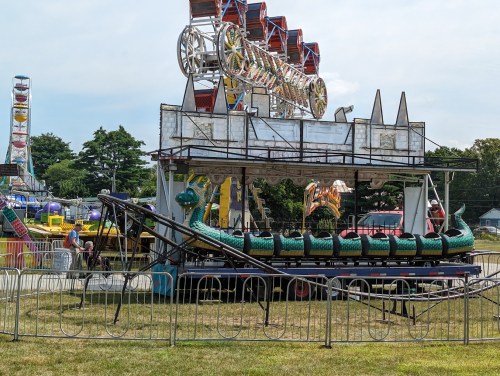 A carnival ride resembling a green caterpillar sits on a platform, surrounded by safety barriers and colorful attractions in the background.