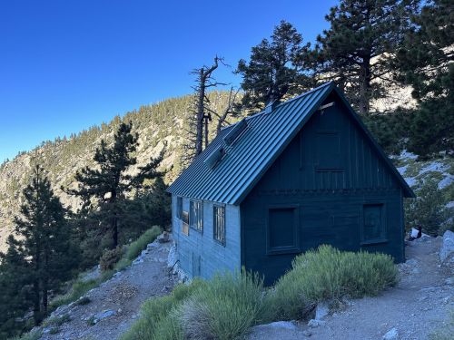 A rustic green cabin nestled among trees on a mountainside, with a clear blue sky in the background.