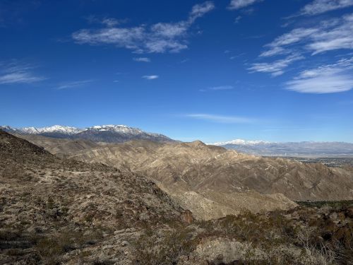 A panoramic view of rugged mountains with snow-capped peaks under a clear blue sky and scattered clouds.