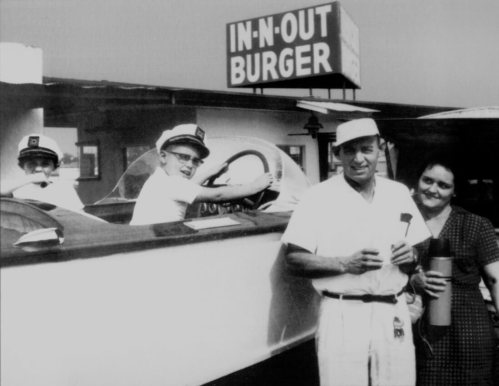 A vintage black-and-white photo of people at an In-N-Out Burger, with staff in uniforms and a couple holding drinks.
