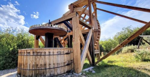 A wooden water wheel structure beside a circular wooden basin, surrounded by greenery and blue sky.