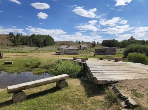 A rustic landscape featuring a wooden bridge, grassy fields, and old buildings under a blue sky with scattered clouds.