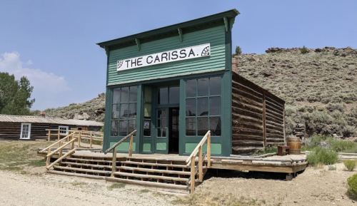 A green wooden building with "The Carissa" sign, surrounded by hills and a dirt path.