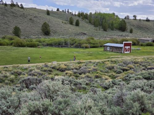 A grassy area with a few people, a red barn, and green hills in the background under a partly cloudy sky.