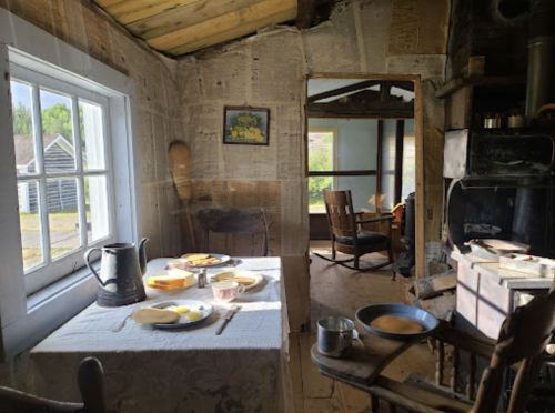 A rustic room with a table set for breakfast, featuring dishes and a kettle, surrounded by newspaper-covered walls.