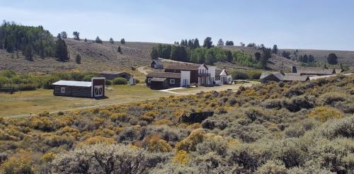 A scenic view of a historic western town with wooden buildings surrounded by sagebrush and rolling hills.