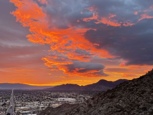 A vibrant sunset with orange and pink clouds over a city and mountains, viewed from a rocky hillside.