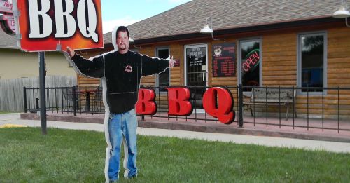 A life-sized cutout of a man holding a BBQ sign stands outside a BBQ restaurant with an "Open" sign in the window.
