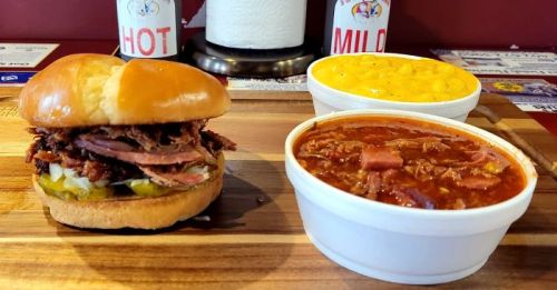 A sandwich with pulled meat and pickles, served with two sides: macaroni and cheese and chili in white bowls.