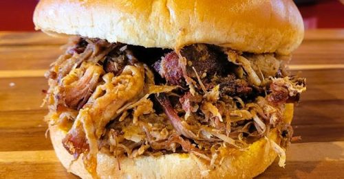 Close-up of a pulled pork sandwich with tender meat piled high in a soft bun, resting on a wooden surface.