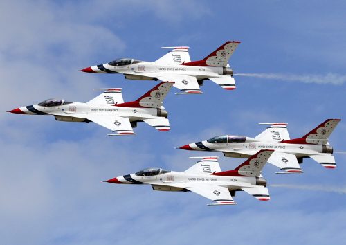 Four F-16 fighter jets in formation, performing a flyover against a blue sky with clouds.