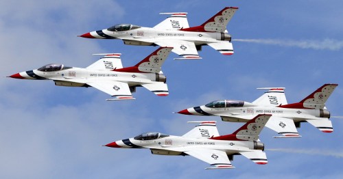 Four F-16 fighter jets in formation, performing a flyover with smoke trails against a blue sky.