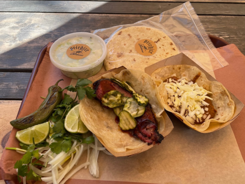 Two tacos with grilled meat and toppings, a side of guacamole, lime wedges, and fresh herbs on a tray.