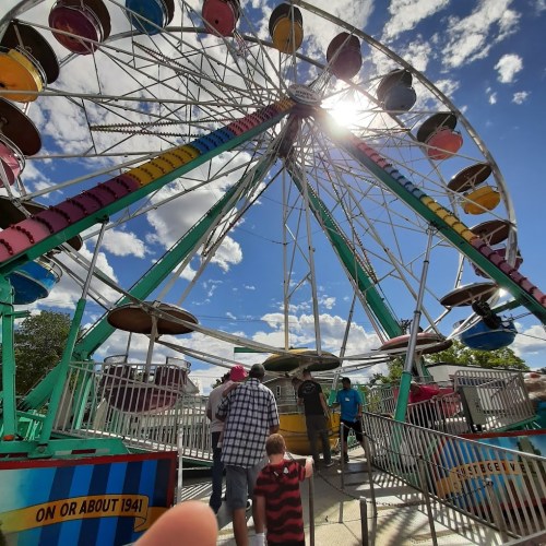 A colorful Ferris wheel with people waiting in line under a bright sky with fluffy clouds.