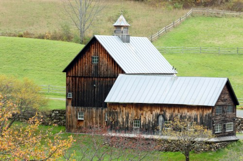 A rustic wooden barn with a metal roof, surrounded by green fields and trees in a serene rural landscape.