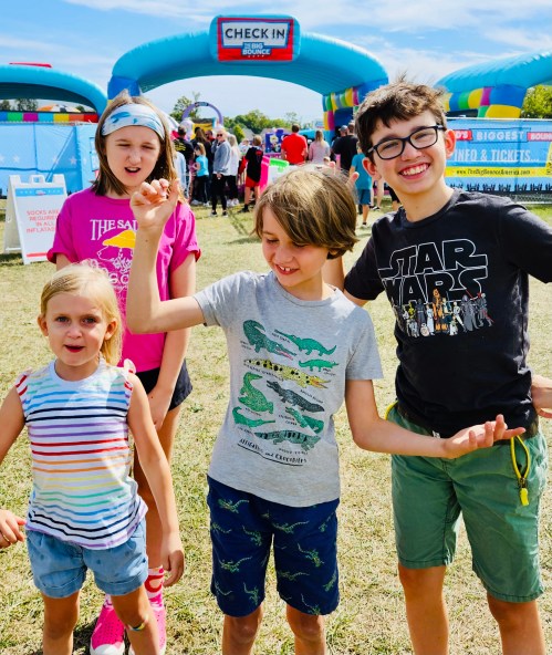 Four children pose playfully in front of a colorful inflatable structure at an outdoor event.