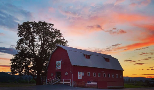A red barn stands under a colorful sunset, with a large tree nearby and mountains in the background.