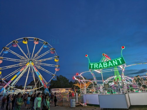 A vibrant carnival scene at dusk featuring a Ferris wheel and a neon-lit ride named "TRABANT."