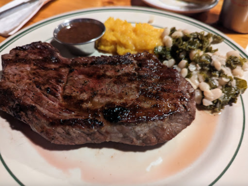 A grilled steak on a plate, accompanied by a small cup of sauce, cornbread, and a side of beans and greens.