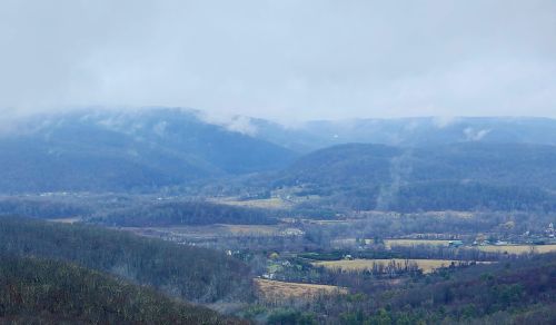 A misty landscape of rolling hills and valleys, with patches of farmland and scattered trees under a cloudy sky.