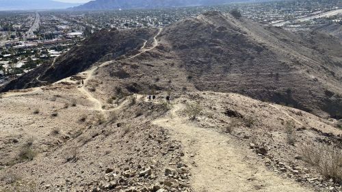A rocky trail winds through a desert landscape, with hikers visible in the distance and mountains in the background.