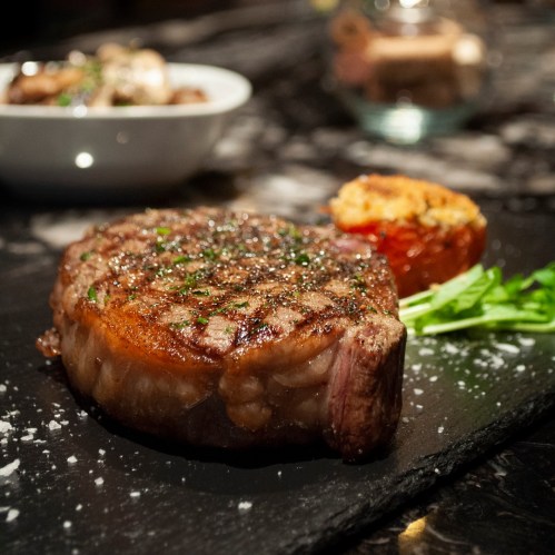 A perfectly cooked steak on a black slate plate, garnished with herbs, alongside a stuffed tomato and greens.