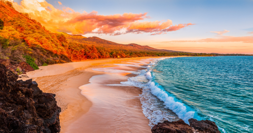 A serene beach at sunset, with golden sand, gentle waves, and vibrant clouds over a lush, green hillside.