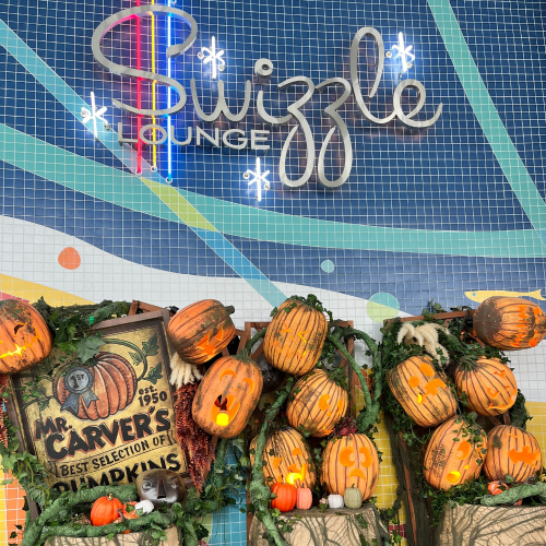 Sign for "Swizzle Lounge" with decorative pumpkins and foliage in a colorful setting.