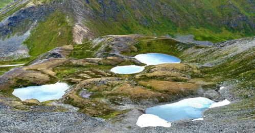 Aerial view of rocky terrain with several small, reflective ponds surrounded by green grass and hills.