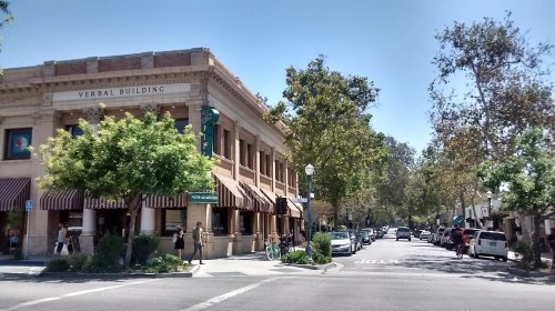 A sunny street scene featuring the Verbal Building, trees, and people walking along the sidewalk.