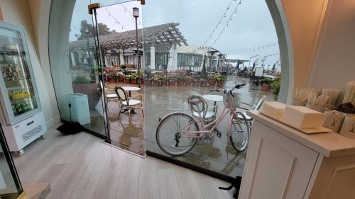 A view from inside a café showing a rainy outdoor scene with a bicycle parked outside and tables under a covered area.