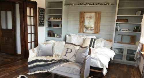 Cozy living room with a sofa, decorative pillows, and shelves filled with books and decor. Warm, inviting atmosphere.