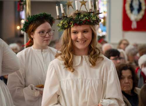 A girl in a white dress and crown of candles smiles while holding a plate, with another girl in the background.