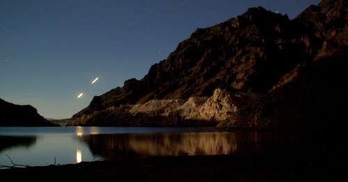 A serene night landscape featuring mountains reflected in calm water under a starry sky.