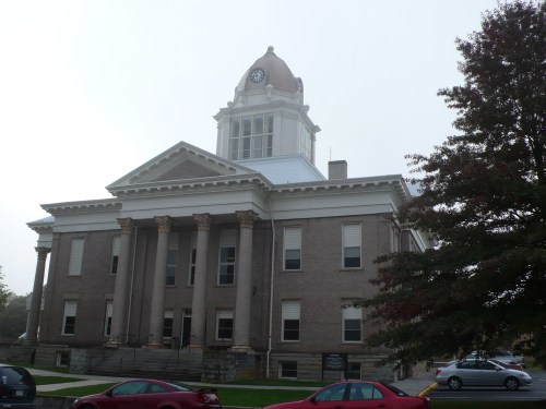 Historic brick building with a clock tower, featuring columns and a foggy background. Red cars parked in front.