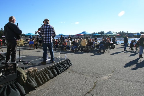 A crowd enjoys a live music performance outdoors by the water, with colorful umbrellas in the background.