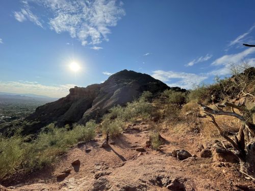 A sunlit mountain peak rises above a rocky trail, surrounded by sparse vegetation and a clear blue sky.