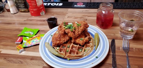 A plate of fried chicken on a waffle, garnished with green onions, pickles, and accompanied by a drink and candy.