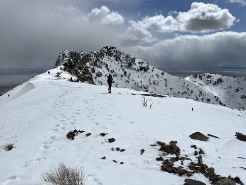 A lone hiker stands on a snowy mountain peak, surrounded by clouds and rocky terrain.