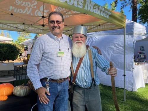 Two men pose together at an outdoor event, one wearing a metal pot as a hat and holding a walking stick.