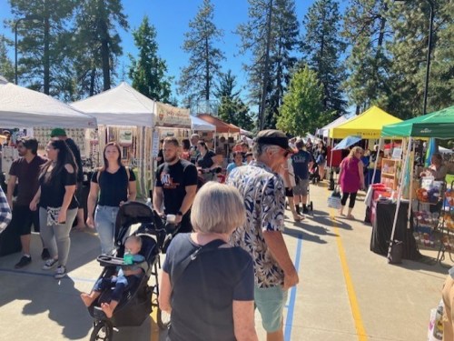A bustling outdoor market with vendors, shoppers, and colorful tents under a clear blue sky and tall trees.