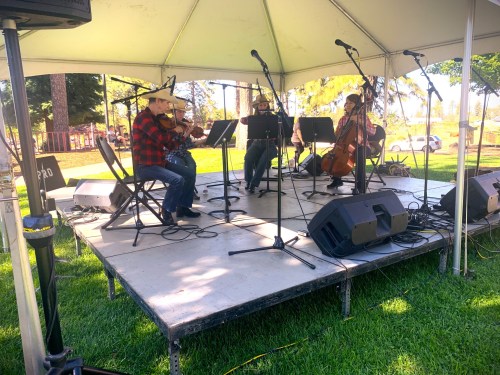 A trio of musicians perform under a tent, playing violin, cello, and guitar on a grassy stage.