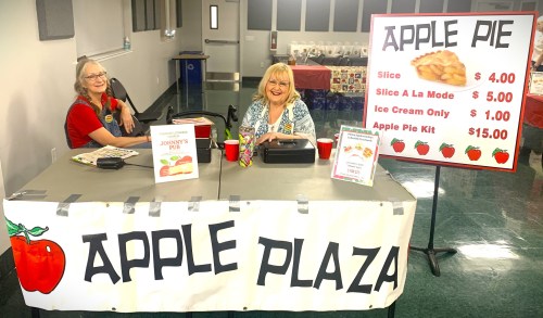 Two women smile behind a table at a fair, selling apple pie and related items, with a large sign displaying prices.