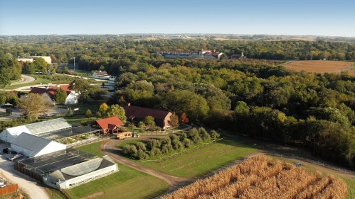 Aerial view of a rural landscape featuring farms, greenhouses, and dense trees under a clear blue sky.
