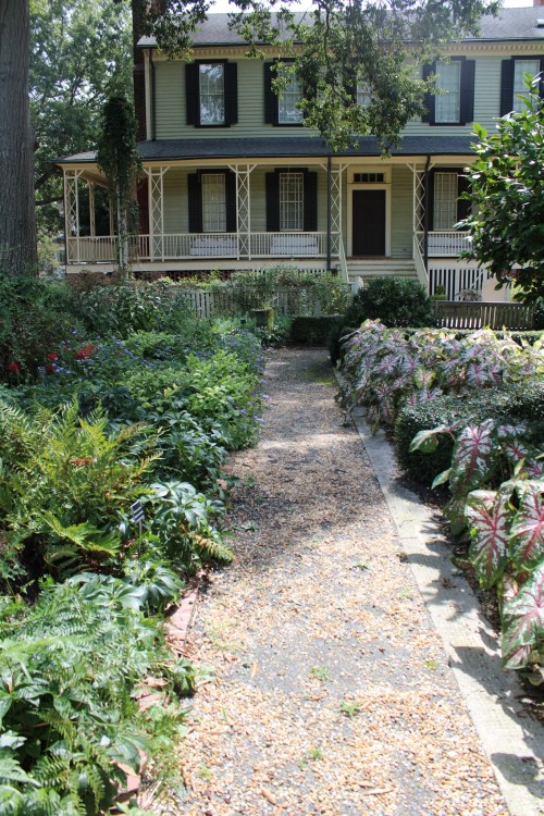 A pathway lined with greenery leads to a two-story house with a porch and large windows, surrounded by a lush garden.