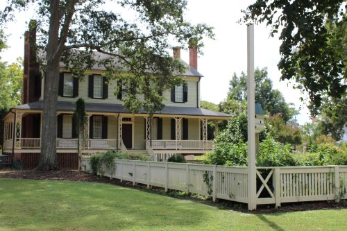 A large, two-story house with a porch, surrounded by greenery and a white picket fence on a sunny day.