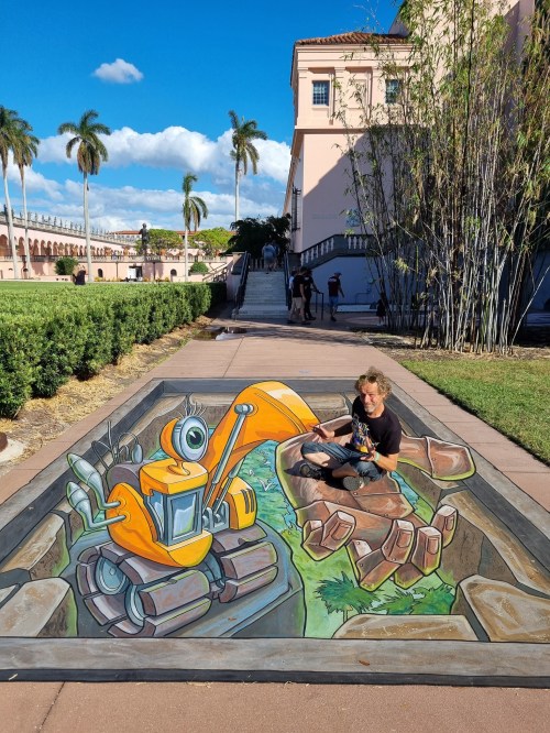 A child sits beside a 3D street art mural of an orange excavator emerging from a rocky pit in a park setting.